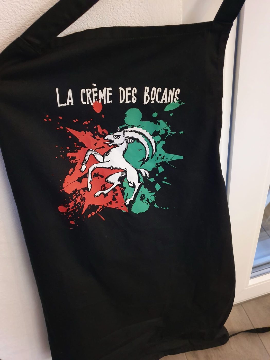 Le tablier noir La crème des Bocans, avec le bouc blanc sur des éclaboussures rouge et vert