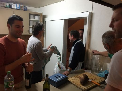Quatre personnes dans une cuisine communale s'affairant autour d'une table avec bouteilles et sacs.