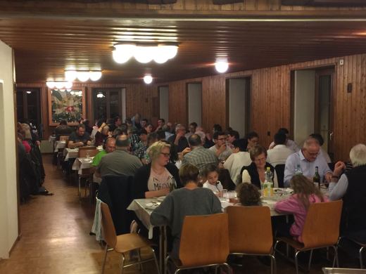 Vue d'ensemble d'une salle communale pleine de convives assis a de longues tables sous eclairage chaud.