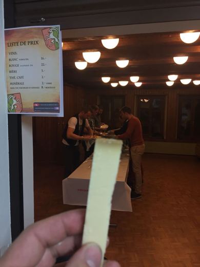 Une tranche de gruyère tenue devant la liste des prix de la soirée, la salle communale et l'équipe en fond