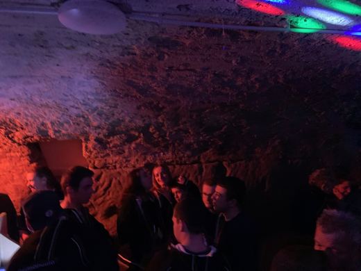 Vue intérieure d'une cave voûtée en pierre éclairée de lumières disco multicolores, avec des silhouettes de personnes en arrière-plan.
