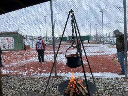 Marmite noire posee sur un trepied au dessus d'un feu de bois, en exterieur sur sol enneige.