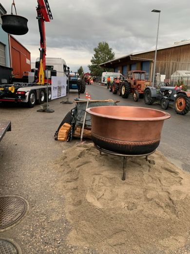Chaudron de cuivre posé sur un tas de sable, camion-grue et tracteurs anciens garés en arrière-plan