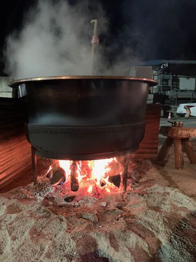 Chaudron noir en pleine ébullition avec des flammes vives dessous, un bras tenant une spatule au-dessus de la vapeur