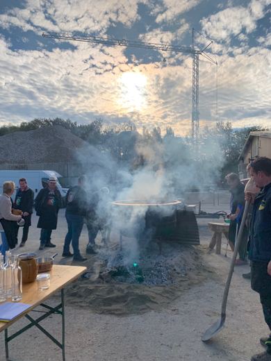 Groupe de personnes rassemblées autour d'un chaudron fumant au crépuscule, grue de chantier visible au loin