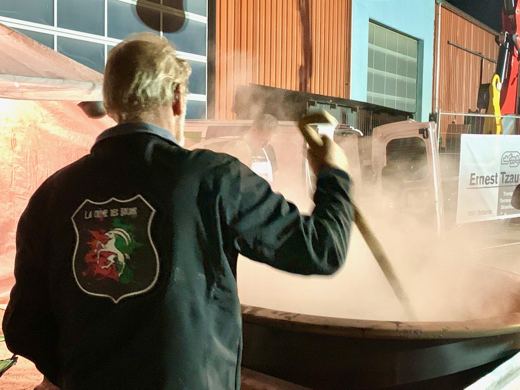 Un homme vu de dos, veste marquée d'un écusson, remuant un chaudron dans un nuage de vapeur