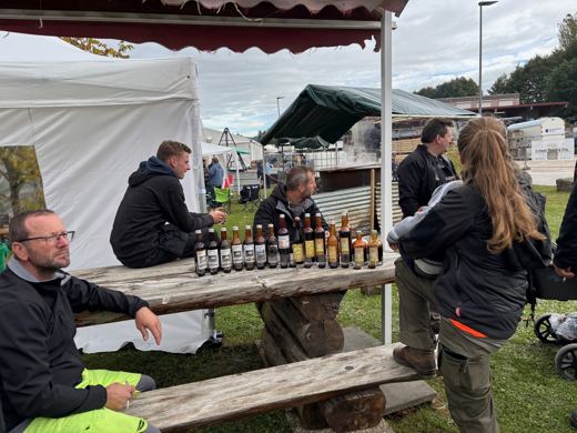 Plusieurs personnes assises et debout autour d'une table à pique-nique avec des bouteilles de raisinée, sous un abri