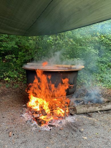 Chaudron noir sous une bâche verte avec de hautes flammes orangées, planche en bois posée sur le dessus