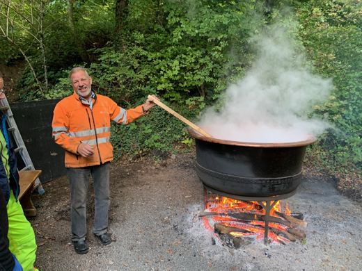 Un homme en veste de sécurité orange pose près d'un chaudron fumant posé sur un feu de bois
