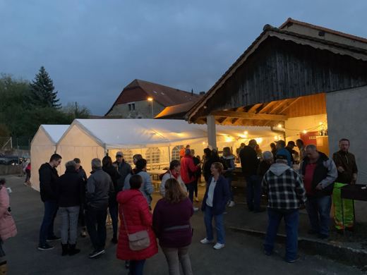 Groupe de personnes rassemblées devant une grange illuminée au crépuscule, tente blanche à gauche