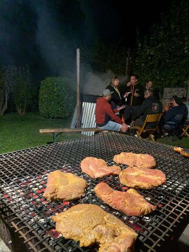 Viande grillant sur des braises au premier plan, un groupe de personnes assises près d'un chaudron fumant à l'arrière-plan