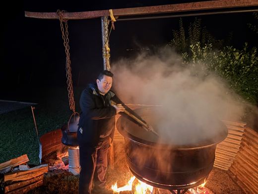 Un homme remue le contenu d'un grand chaudron fumant avec une longue spatule en bois, de nuit