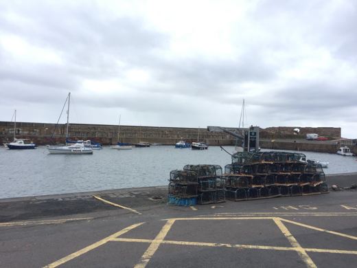 Petit port écossais avec voiliers amarrés et casiers de pêche empilés sur le quai, ciel nuageux.
