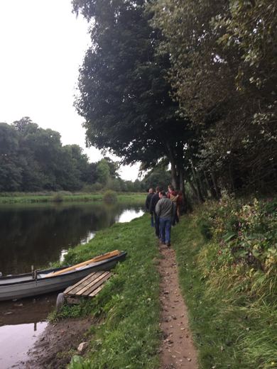 Sentier boisé longeant une rivière calme, une personne marchant au loin, petite barque amarrée.