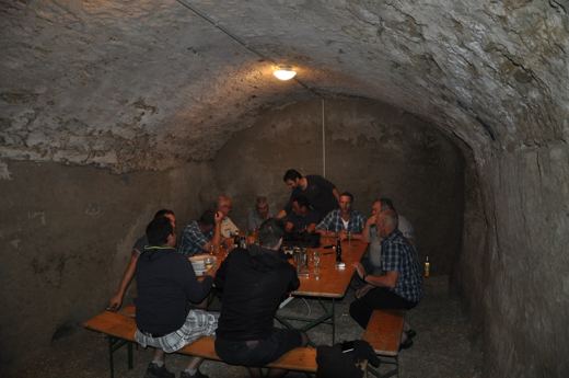 Groupe de personnes assises à une longue table dans une cave en pierre voûtée, verres et bouteilles sur la table.