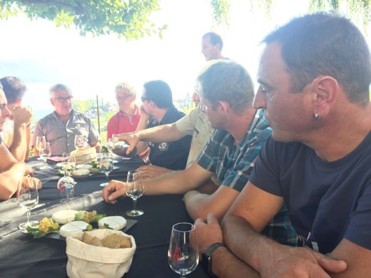 Groupe de convives assis autour d'une table dressée en plein air, verres de vin et plats posés devant eux.