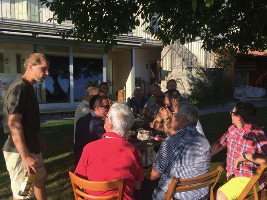 Un homme debout s'adresse à un groupe assis autour de tables de jardin devant un bâtiment.