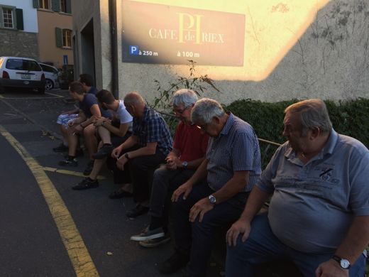 Groupe d'hommes assis à l'ombre sur un muret devant un café de village, voitures garées à proximité.