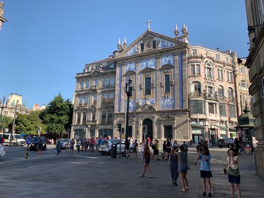 Grande place pavée avec passants et façade richement ornée d'un édifice historique portugais, ciel bleu.