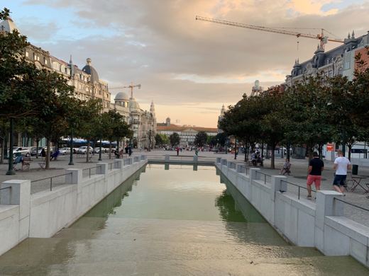 Canal rectiligne servant de miroir d'eau au centre d'une place, bâtiments en fond, lumière dorée du soir.