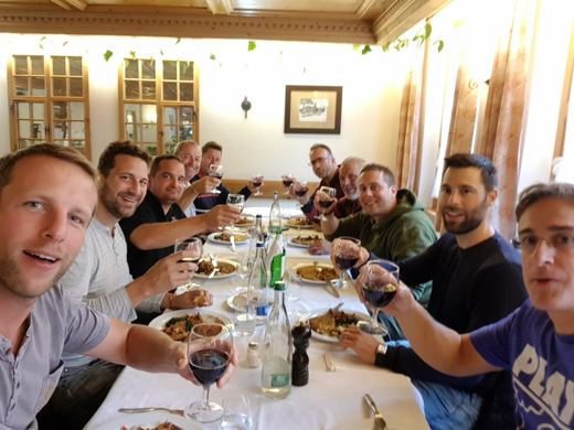 Groupe d'hommes attablés dans une salle à manger, verres de vin levés pour un toast, plats sur la table.