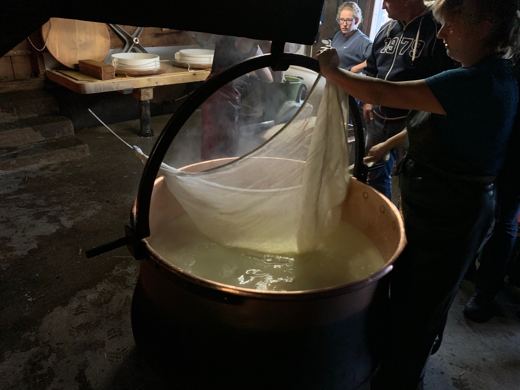Une personne soulève un linge rempli de caillé hors d'un grand chaudron de cuivre, spectateurs en arrière-plan.