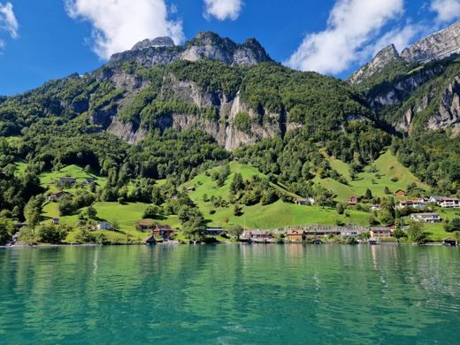 Vue panoramique d'un village suisse au bord d'un lac turquoise, entouré de montagnes verdoyantes sous un ciel nuageux.