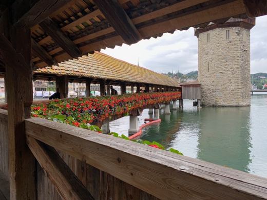 Pont couvert en bois orné de bacs à fleurs, tour d'eau octogonale en arrière-plan, rivière calme à Lucerne.
