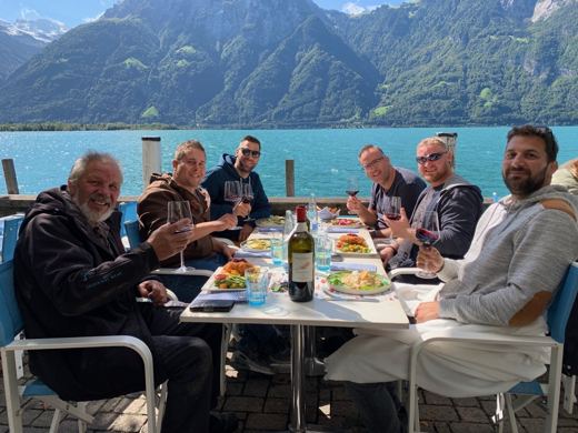 Groupe d'hommes attablés en terrasse ombragée au bord d'un lac turquoise, repas et boissons sur la table.