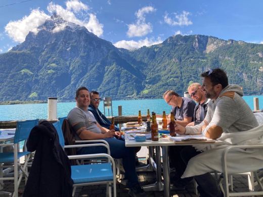 Groupe d'hommes attablés en terrasse extérieure, lac et sommets enneigés en arrière-plan, repas en cours.