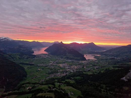 Coucher de soleil aux couleurs rouge orangé sur une vallée et un lac, silhouettes de montagnes au loin.