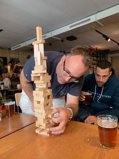 Deux hommes concentrés autour d'une tour de Jenga en bois très haute, verres de bière sur la table.