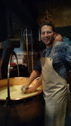 Un homme souriant tire le caillé hors d'une chaudière de cuivre, tablier de fromager sur les épaules.