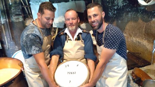 Trois hommes souriants posent derrière une meule de fromage fraîchement pressée, dans une fromagerie traditionnelle.