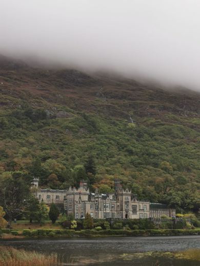 Grand édifice de style abbaye au bord d'un lac, entouré de collines boisées dans la brume.