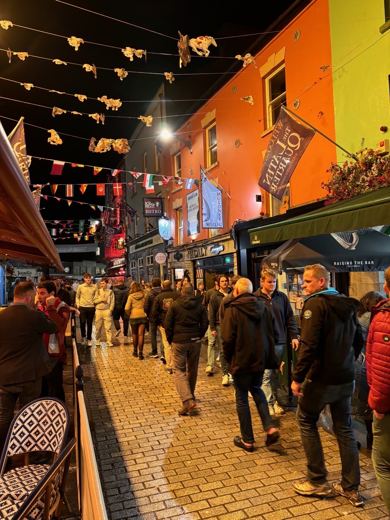 Rue piétonne animée bordée de façades colorées, guirlandes lumineuses tendues au dessus, passants en soirée.
