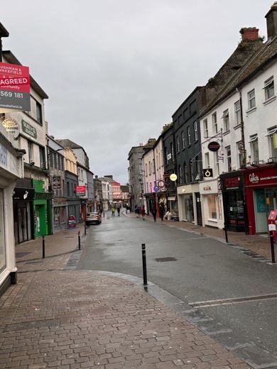 Rue commerçante bordée de façades colorées, vide de passants, sous un ciel gris et couvert.
