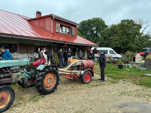 Groupe de personnes rassemblées devant une ferme au toit rouge, vieux tracteur agricole garé à côté, minibus en arrière-plan.