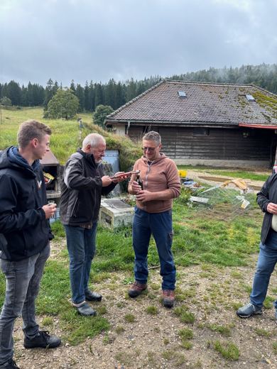 Trois hommes debout dans une cour de ferme, l'un tendant une bouteille à un autre, bâtiments anciens en arrière-plan.