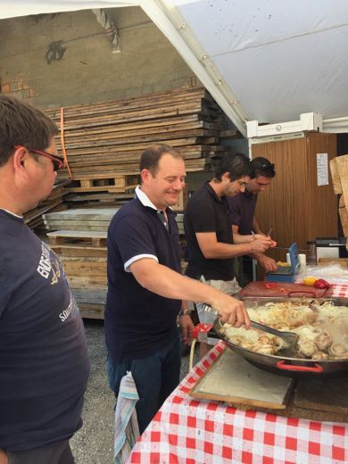 Deux hommes souriants se tiennent près d'une grande poêle sous une tente, lors d'une préparation culinaire en plein air.